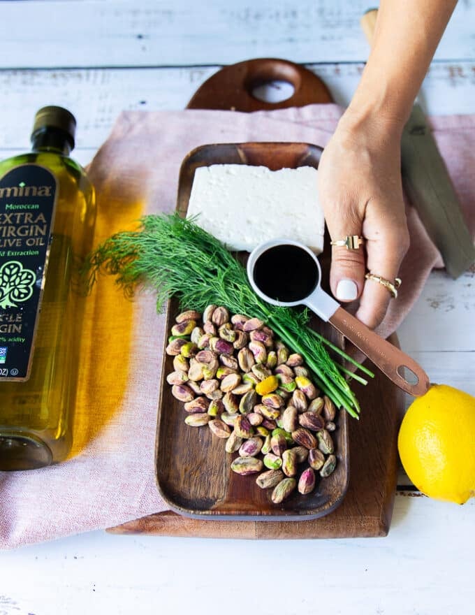 Ingredients for carpaccio topping including a board with some feta, a bowl of pistachios, some dill leaves, a spoon of balsamic syrup, one lemon, and a bottle of olive oil