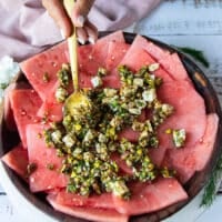 The watermelon slices placed on a wooden plate and a hand spooning some of the carpaccio topping with a spoon over the watermelon slices