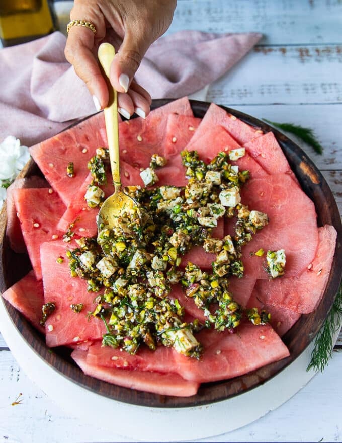 The watermelon slices placed on a wooden plate and a hand spooning some of the carpaccio topping with a spoon over the watermelon slices