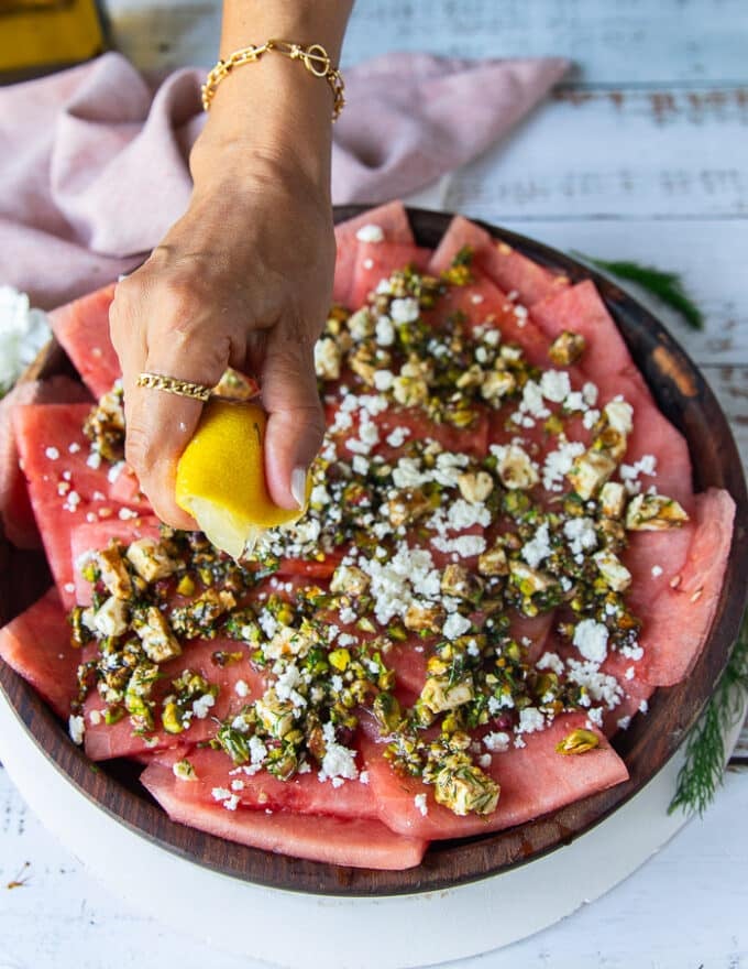 A hand squeezing lemon over the watermelon carpaccio over the topping to finish off the carpaccio recipe