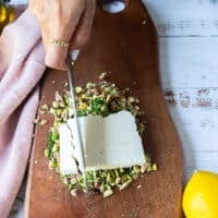 a block of feta placed over the chopped pistachio and dill and a hand holding a knife to chop the feta too