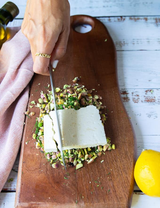 a block of feta placed over the chopped pistachio and dill and a hand holding a knife to chop the feta too