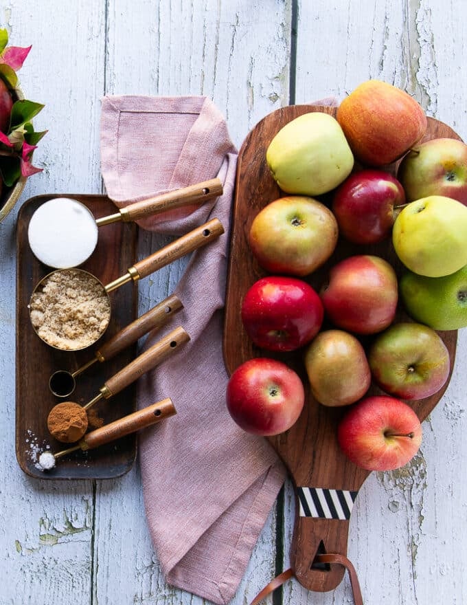 ingredients to make apple butter recipe including apples in a wooden bowl, some white sugar in a cup, brown sugar in a cup, cinnamon, salt and vanilla in separate spoons
