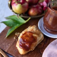 A croissant spread with apple butter on a wooden board surrounded by a bowl of apples and a jar of homemade apple butter