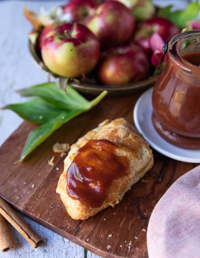 A croissant spread with apple butter on a wooden board surrounded by a bowl of apples and a jar of homemade apple butter