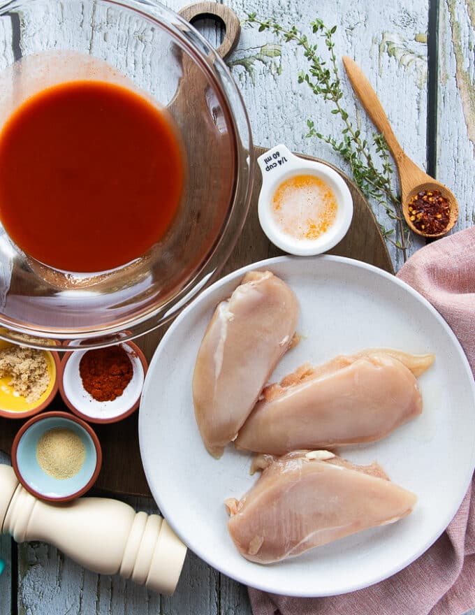 ingredients on a table for making buffalo chicken wraps. Including: a plate of chicken breasts, a bowl of hot sauce, a small bowl of meted butter, small bowls of spice like garlic powder, salt, pepper, brown sugar, chilli flakes and paprika
