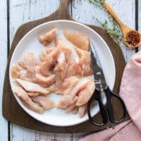 Chicken breasts cut up into strips on the plate using scissors shown next to the plate