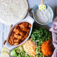 All ingredients ready to assemble the buffalo chicken wrap on a board. Including a bowl of blue cheese sauce, the cooked buffalo chicken in a bowl, shredded carrots, diced celery, shredded cheese, sliced avocados, lettuce. green onions and flour tortillas