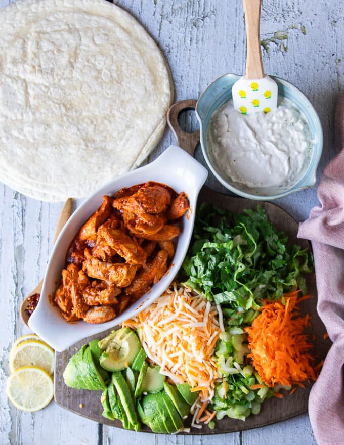 All ingredients ready to assemble the buffalo chicken wrap on a board. Including a bowl of blue cheese sauce, the cooked buffalo chicken in a bowl, shredded carrots, diced celery, shredded cheese, sliced avocados, lettuce. green onions and flour tortillas