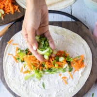 A hand adding lettuce and green onions over the blue cheese spread along the center of a tortilla.