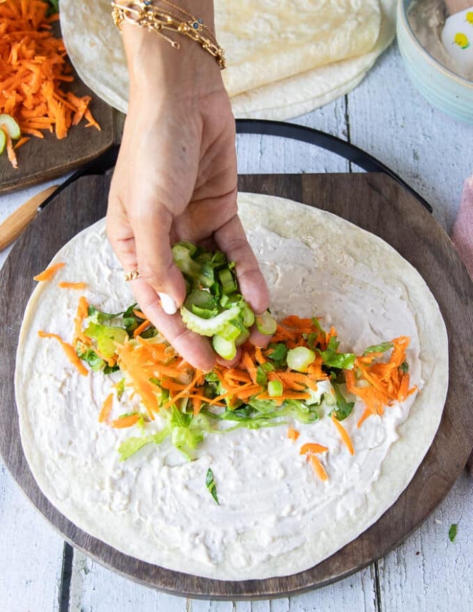 A hand adding lettuce and green onions over the blue cheese spread along the center of a tortilla.