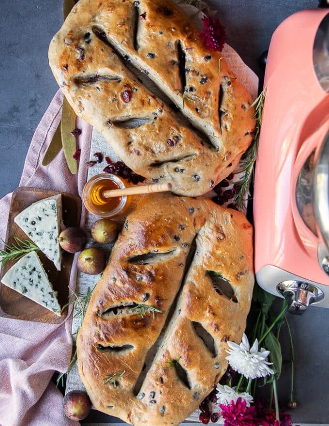 two loaves of baked Fougasse breads on a cheese board surrounded by cheese, fruits and honey.