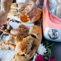 A hand drizzling honey on a loaf of fougasse bread over a cheese board