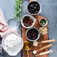 Ingredients in small bowls on a wooden board include bowl of flour, teaspoon of yeast, salt and olive oil in a bowl along with a bowl of cranberries, fresh rosemary, a bowl of olives, some walnuts in a bowl and sun dried tomatoes in a bowl