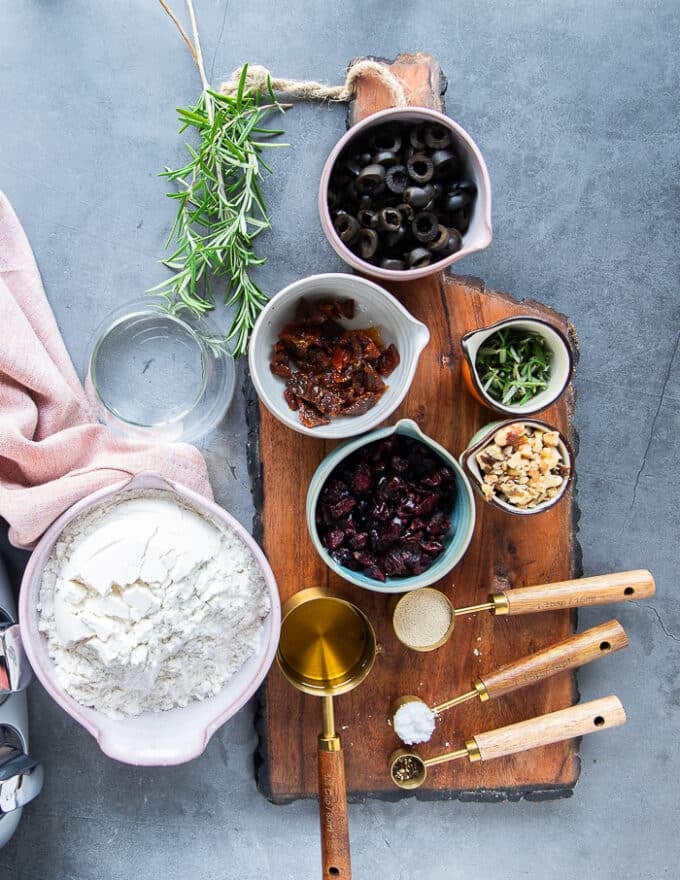 Ingredients in small bowls on a wooden board include bowl of flour, teaspoon of yeast, salt and olive oil in a bowl along with a bowl of cranberries, fresh rosemary, a bowl of olives, some walnuts in a bowl and sun dried tomatoes in a bowl