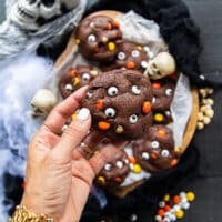 A hand holding one halloween cookie closeup showing the candy eye balls, the chocolatey cookies and the colored chocolate