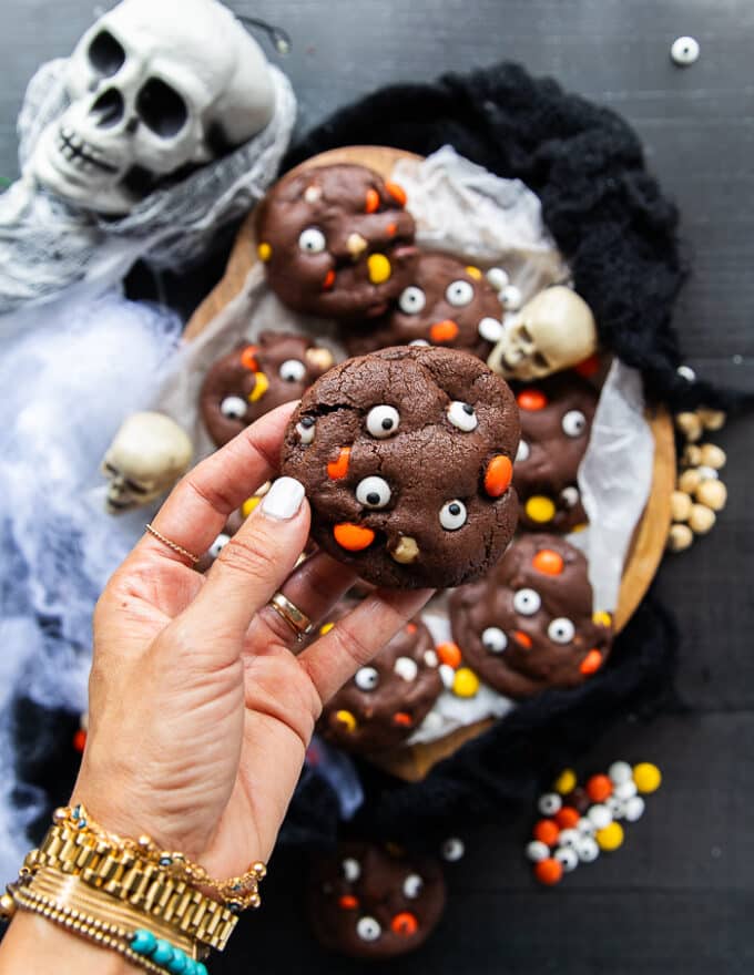 A hand holding one halloween cookie closeup showing the candy eye balls, the chocolatey cookies and the colored chocolate