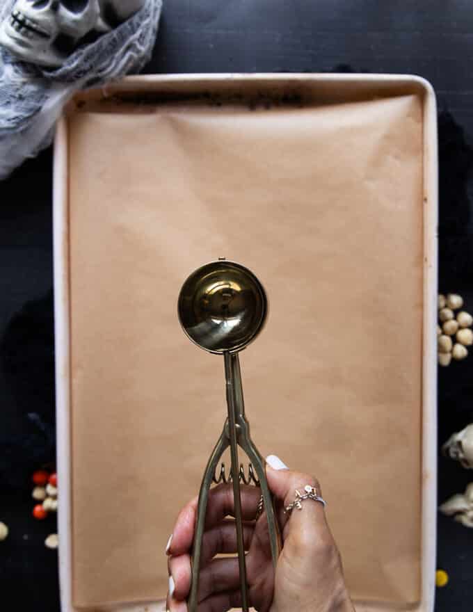 A hand holding a 2 inch ice cream scoop over a baking sheet lined with parchment paper