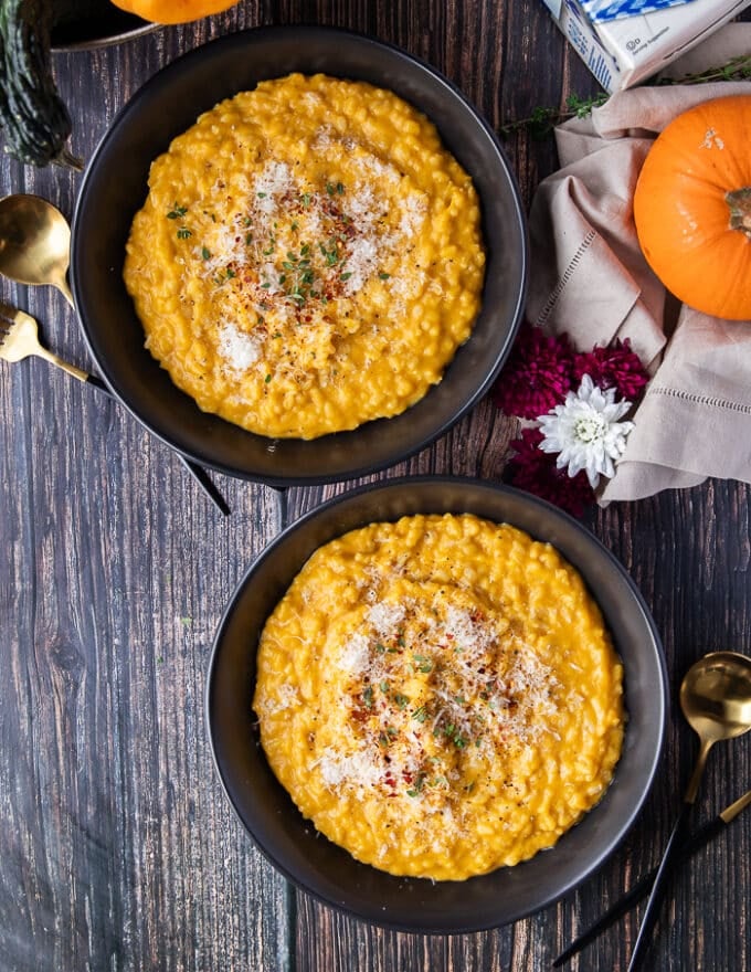 two plates of pumpkin risotto on a wooden table garnished with parmesan cheese and surrounded by fresh baby pumpkins