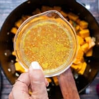 A hand holding stock and ready to pour it into the pot of pumpkin