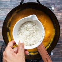 A hand holding a cup of arborio rice and ready to pour it into the pot to make risotto