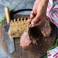 A hand slicing a piece of the oven roasted lamb rack showing the medium rare meat and how tender it is