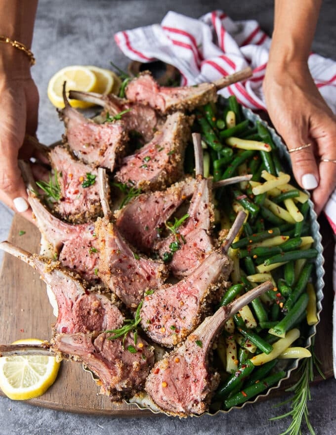 A hand holding a platter of sliced rack of lamb served next to steamed green beans 