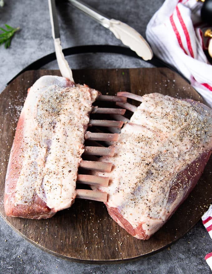 Two French cut racks of lamb on a wooden board seasoned with salt and pepper liberally on both sides