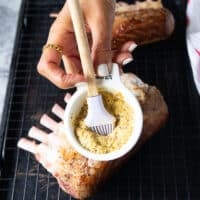 A hand holding a bowl of mustard and a brush to brush the surface of the lamb rack with mustard