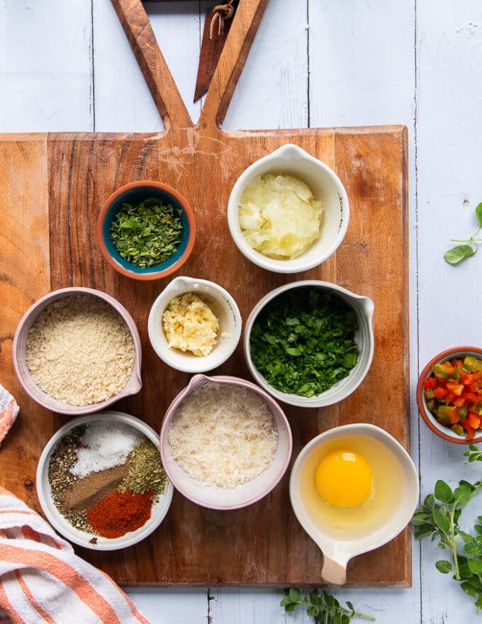 A wooden board with small bowl of ingredients to be added to make the air fryer meatballs. Ingredients include: an egg, spice blend, parsley, panko bread crumbs, parmesan cheese, garlic minced, grated onions