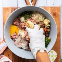 A hand wearing a plastic glove mixing the meat and ingredients together
