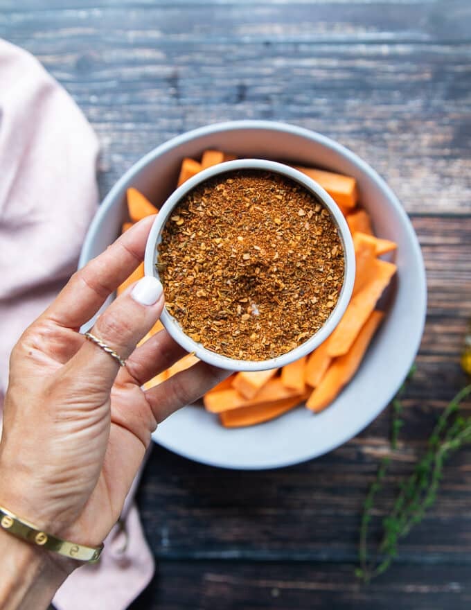 A hand holding a dry spice blend in a plate with a teaspoon of chilli powder, garlic powder, salt, pepper, oregano 