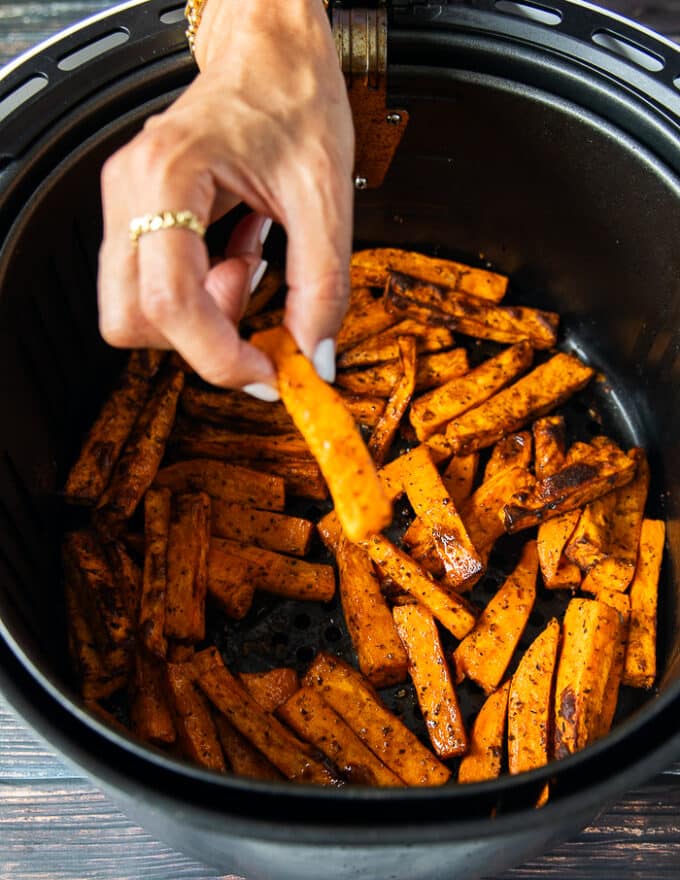 A hand holding one sweet potato fries over the cooked fries in the air fryer showing how crisp it is