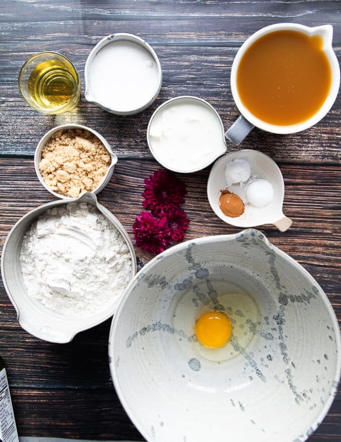 ingredients on a board in small bowls to make apple cider donuts including a bowl of egg, a bowl of flour, baking soda, baking powder, cinnamon, a bowl of brown sugar, a bowl of white sugar, bowl of oil, bowl of yogurt and a large bowl of apple cider