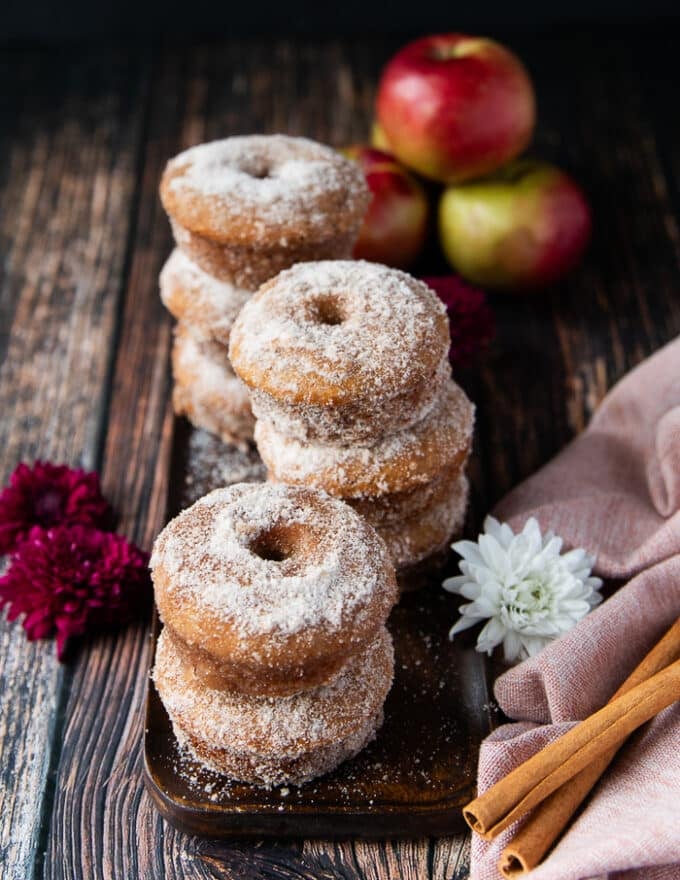 stacks of apple cider donuts surrounded by a stack of apples, a pink kitchen towel and cinnamon sticks