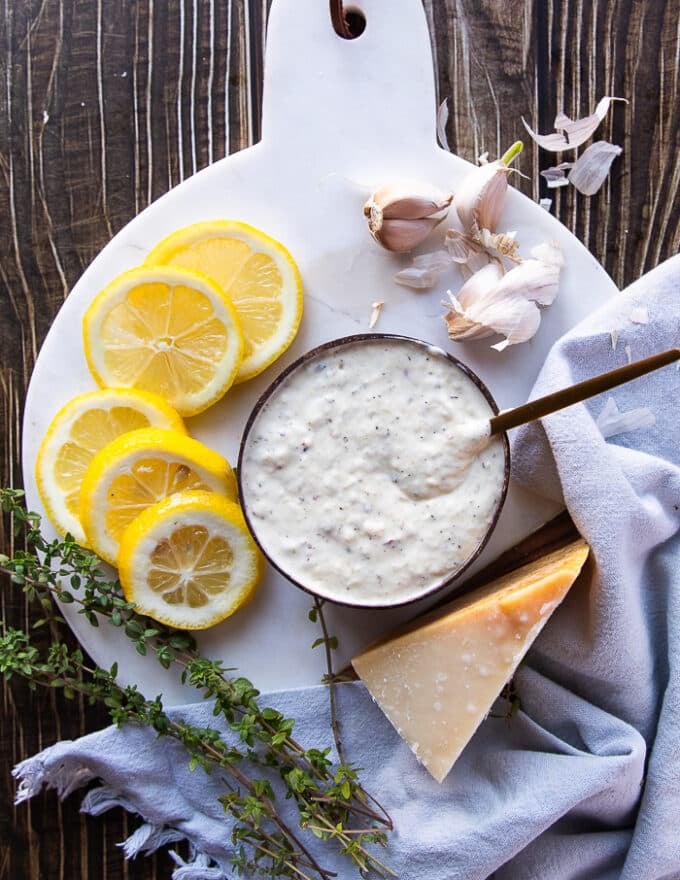 A bowl of garlic parmesan sauce on a white marble surrounded by lemon slices, a wedge of parmesan cheese, fresh thyme leaves and whole garlic cloves