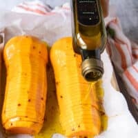 A hand pouring olive oil over seasoned butternut squash halves on a parchment lined baking sheet