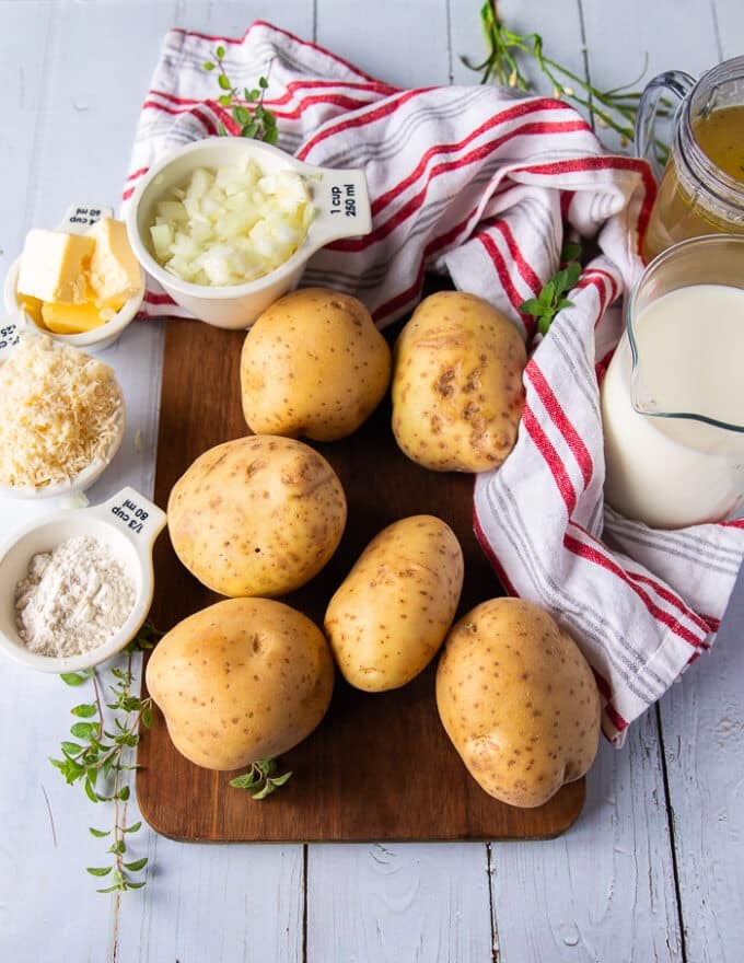 Yellow gold potatoes on a wooden board surrounded by a bowl of flour, a bowl of butter, a bowl of parmesan cheese, some diced onions, some milk in a cup and some stock in a cup