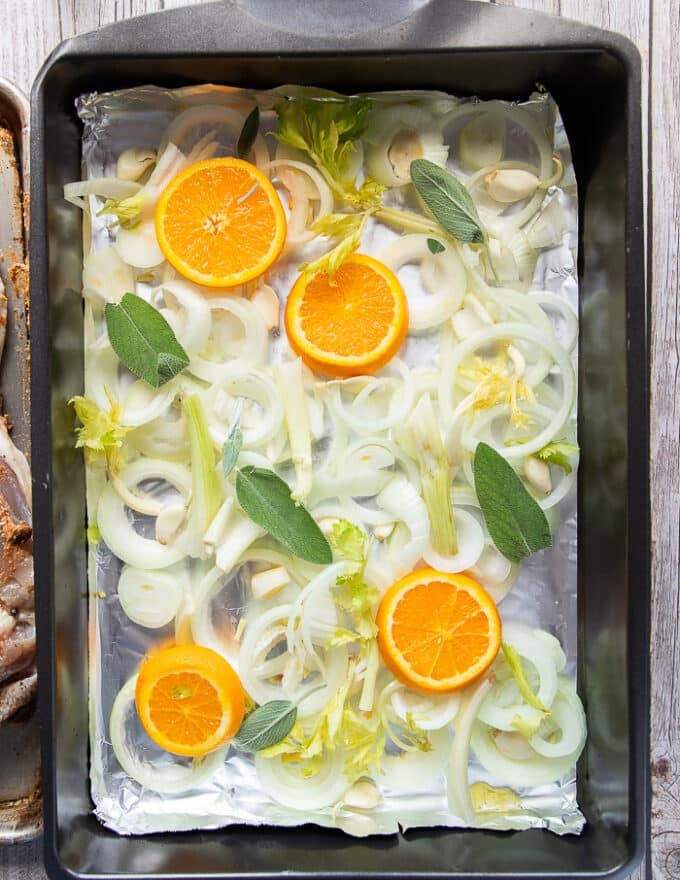 A roasting pan ready and layered with onion rings, orange slices, celery and sage at the bottom