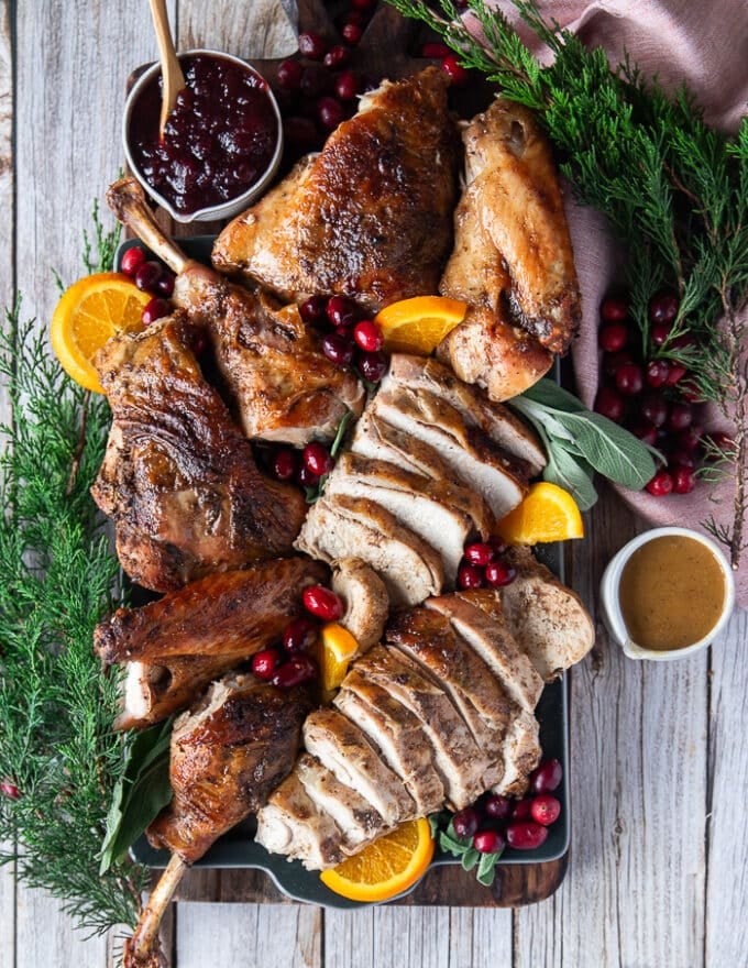 Top view of a whole turkey carved on a platter surrounded by a bowl of cranberries, a bowl of gravy, some orange slices, some fresh cranberries and fresh sage