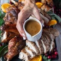 A hand holding a gravy boat made using the pan dripping of the turkey and ready to pour over the craved and sliced spatchcock turkey recipe on the platter