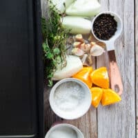 turkey brine ingredients on a table including a large roasting pan with water, a bowl of salt, bowl of sugar, orange slices, quartered onions, whole garlic cloves, fresh herbs, whole peppercorns in a bowl