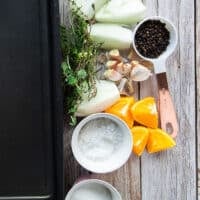 turkey brine ingredients on a table including a large roasting pan with water, a bowl of salt, bowl of sugar, orange slices, quartered onions, whole garlic cloves, fresh herbs, whole peppercorns in a bowl