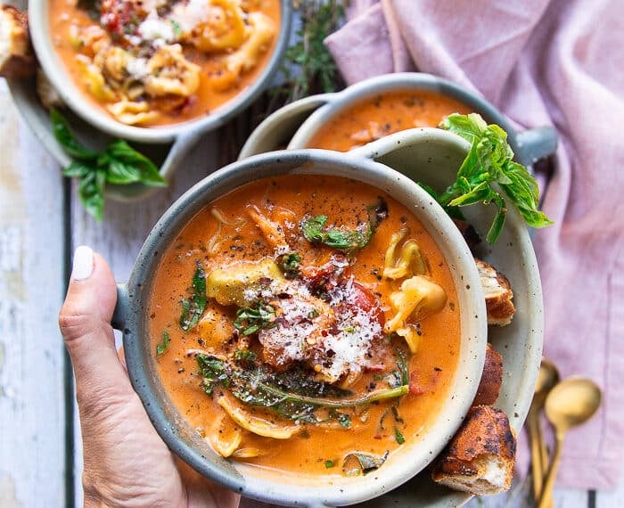 A hand holding one bowl of tortellini soup close up showing the cooked tortellini in a creamy tomato broth with basil leaves and garnished with fresh parmesan cheese