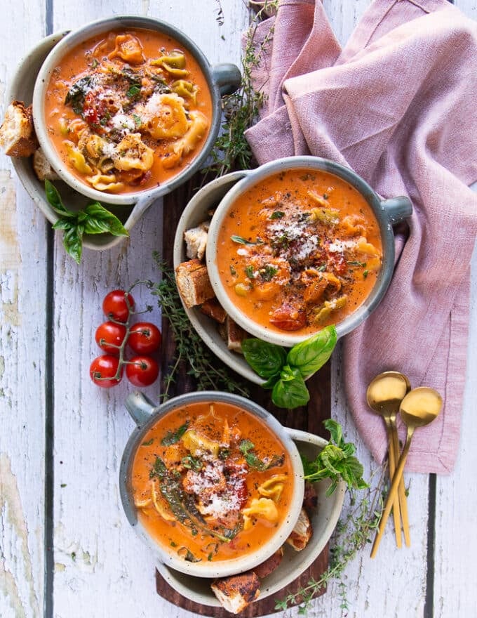 3 bowls of tortellini soup on a wooden board garnished with parmesan cheese on top and surrounded by serving spoons and baby cherry tomatoes, and basil leaves