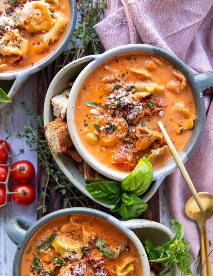 A spoon inside a bowl of creamy tortellini soup surrounded by fresh basil leaves