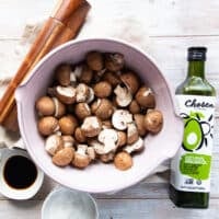 air fryer mushroom ingredients on a board which includes a bowl of mushrooms, a bottle of oil, some salt, pepper and soy sauce