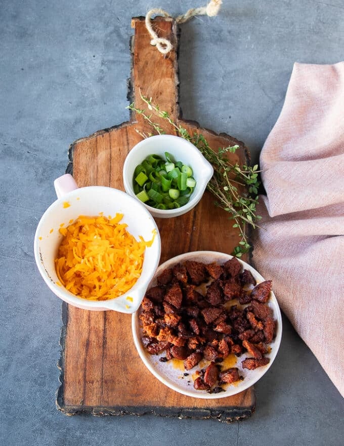 A board with small bowls of topping for the baked soup recipe including the crumbled bacon in a plate, some shredded cheese in a bowl and some green onions minced