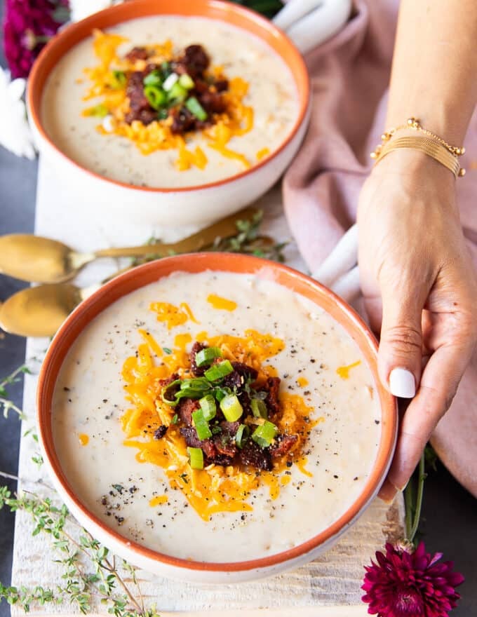 A hand holding a bowl of baked potato soup finished with crumbled bacon or sausage, some green onions and cheddar cheese on top