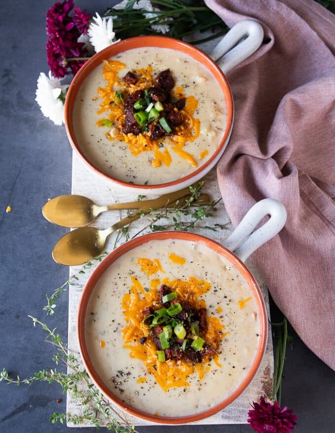 two bowls of baked potato soup on a white board surrounded by a pink kitchen towel and two spoons. Both bowls are garnished with bacon crumbles or sausage crumbles, green onions and cheeses